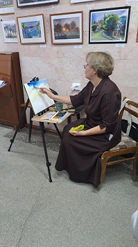 Художница пишет акварель за мольбертом на фоне выставленных картин